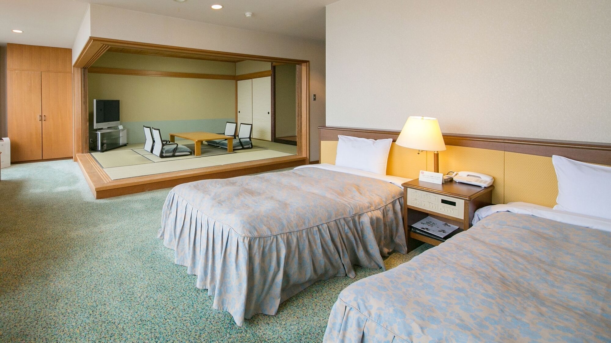 Quarto de hotel com duas camas, uma mesa de cabeceira com abajur, uma pequena mesa com cadeiras e um espelho grande.