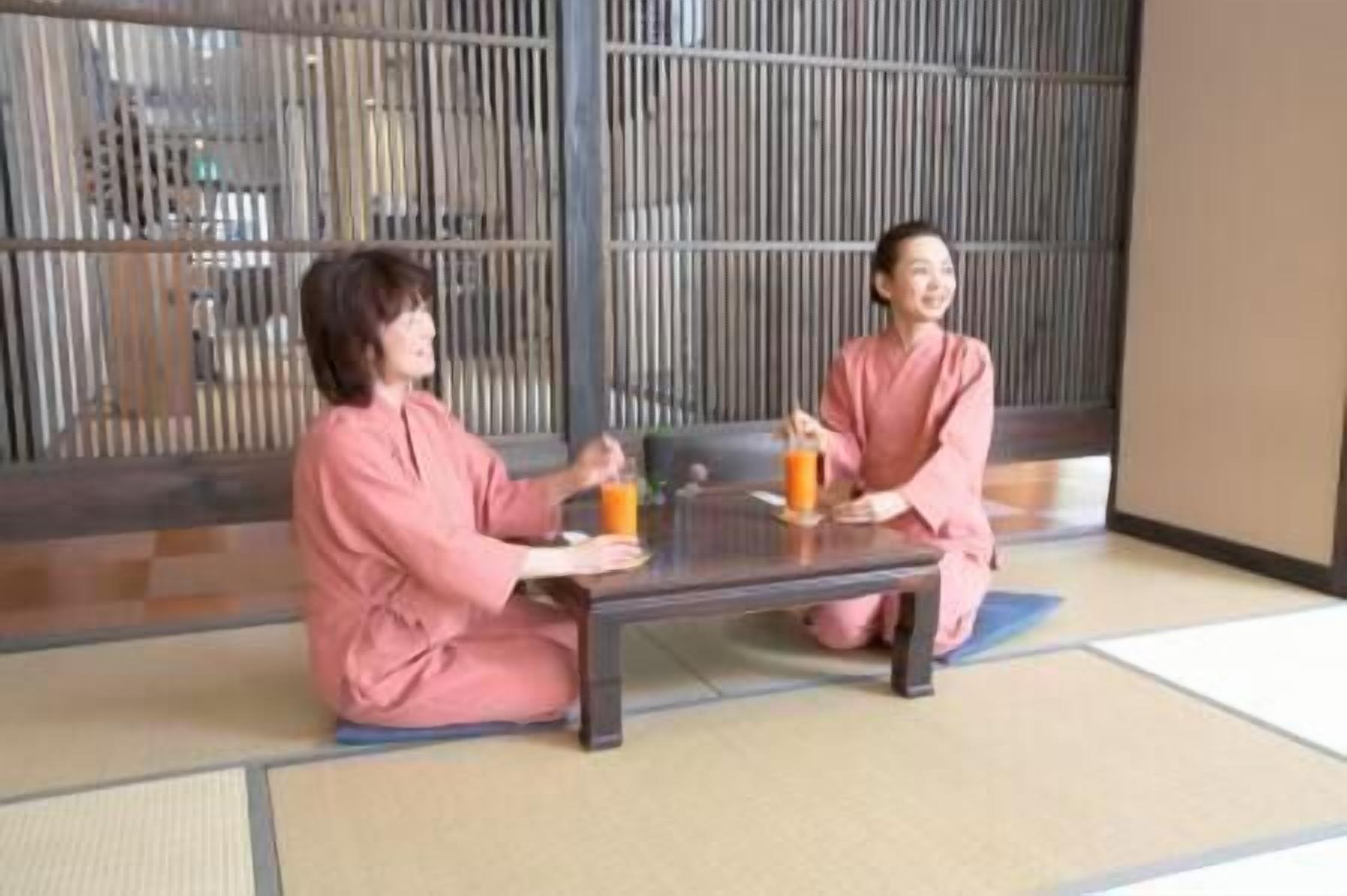 Two individuals in pink robes sitting on tatami mats at a low table with a beverage.