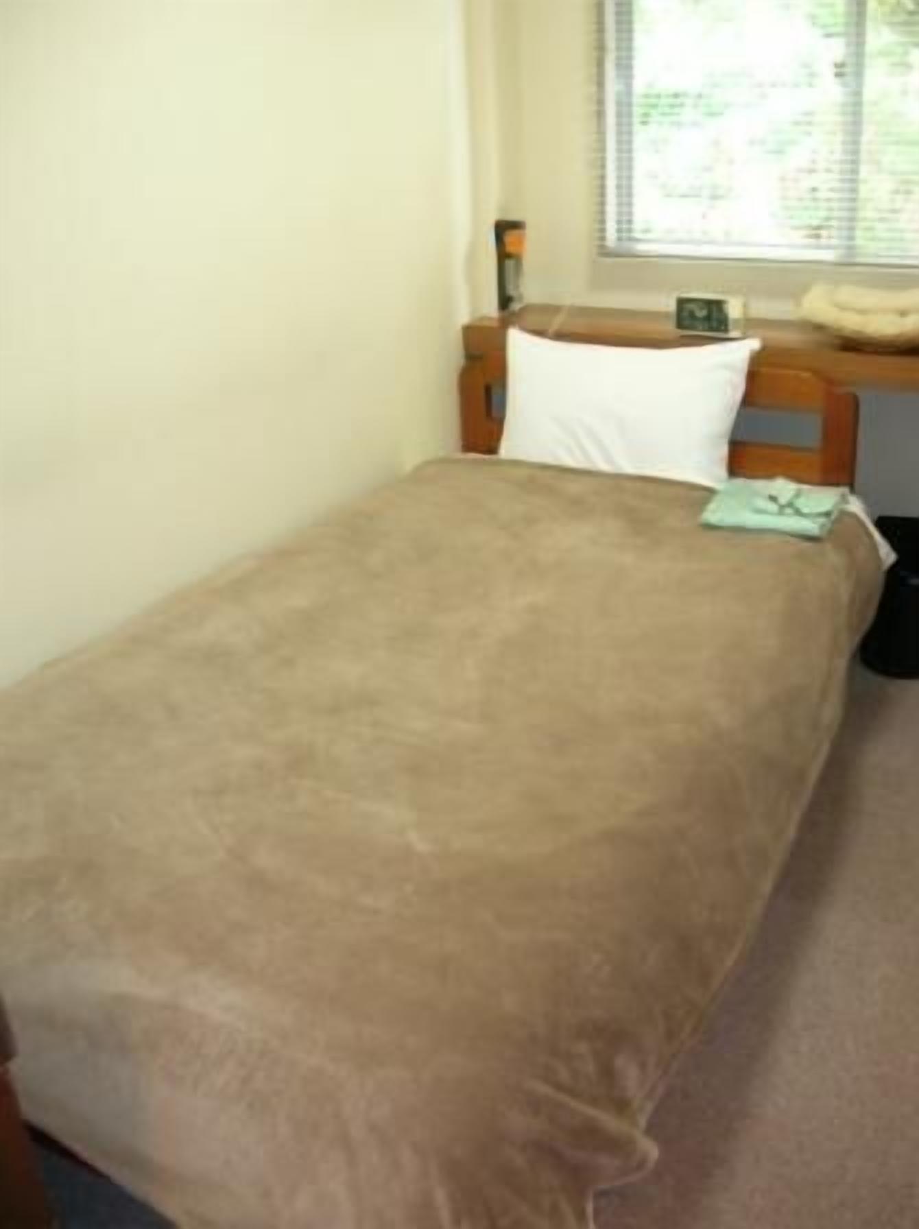 A single bed with a brown blanket, a wooden headboard, a bedside table, and a window with blinds.