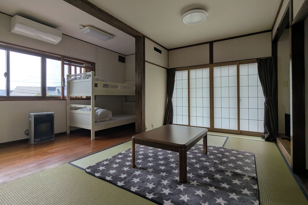 A room with bunk beds, a small table, tatami mats, a heater, and a window with a view.