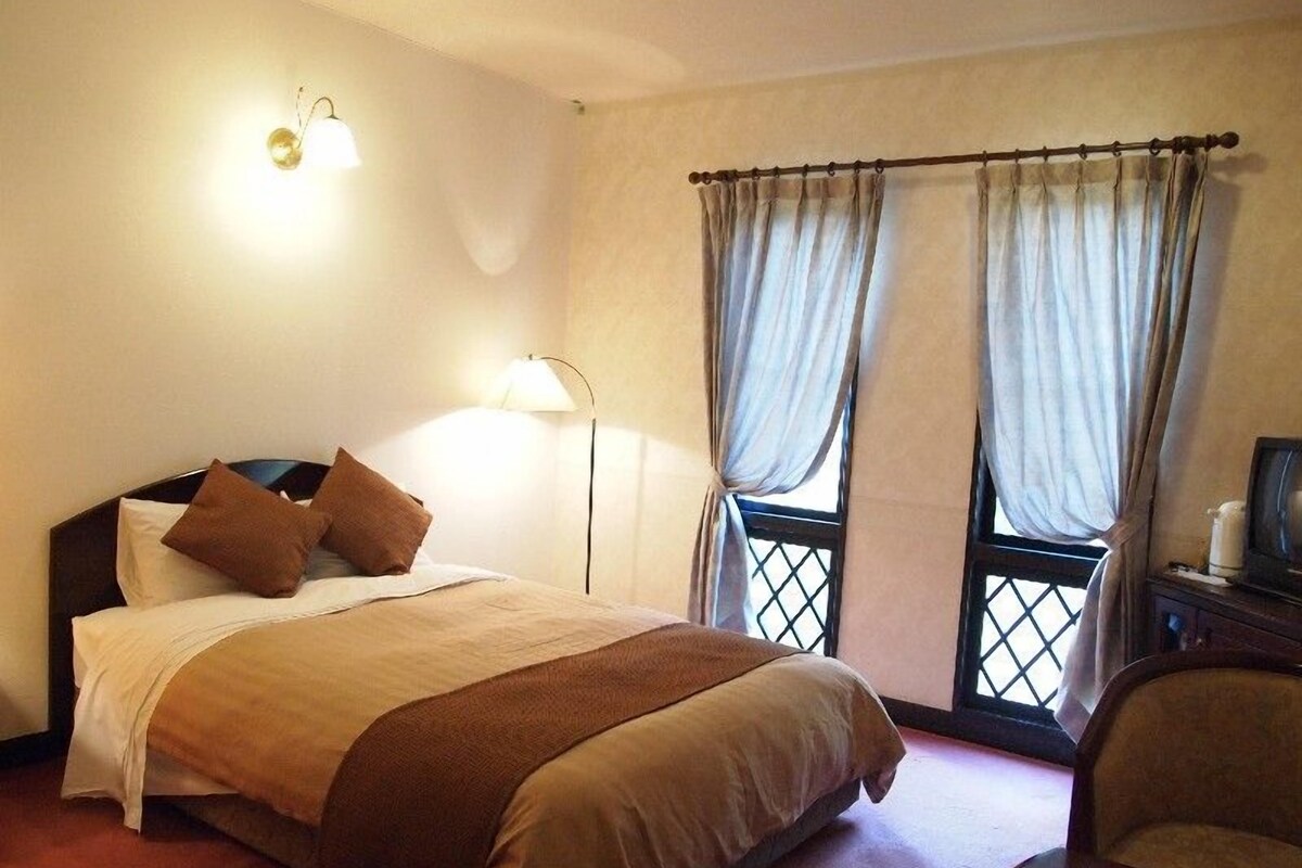 Una camera d'albergo con un letto, due finestre con tende, una lampada da comodino e una sedia.