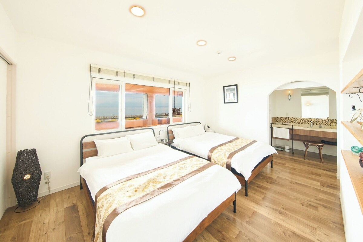 A hotel room with two beds, a wooden floor, a large window with a view, a bathroom with a mirror and sink, and a small table.