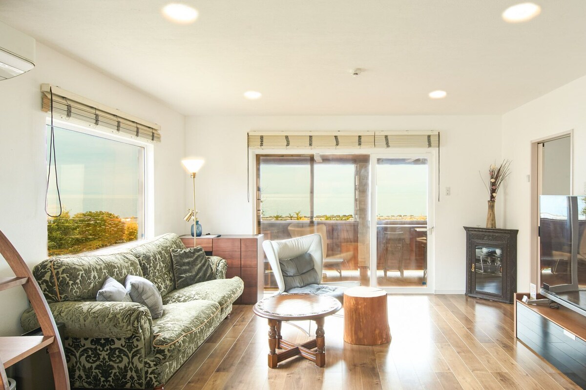 A spacious living room with a sofa, a chair, a round wooden table, and a view through a sliding glass door to an outdoor balcony.