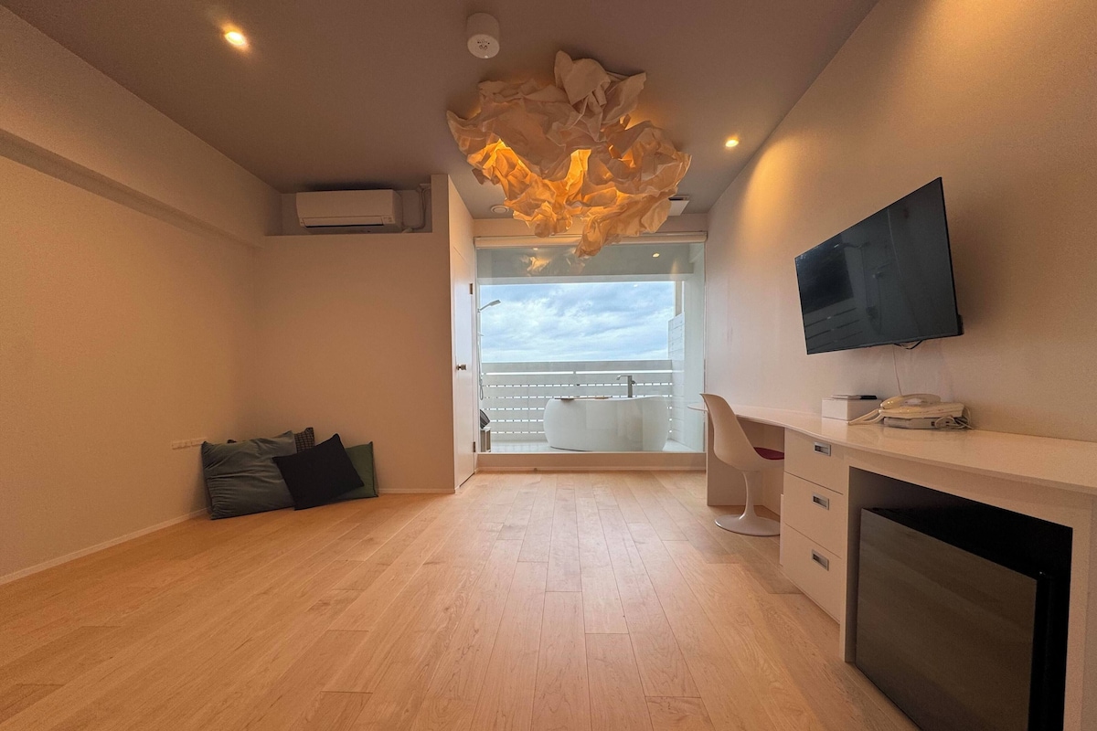 A modern room with a wooden floor, a flat-screen TV, a desk with a telephone, and a view of the sky through a balcony door.