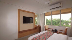 A hotel room with a bed, a TV mounted on the wall, a window with curtains, a chair, and a table with a coffee pot.