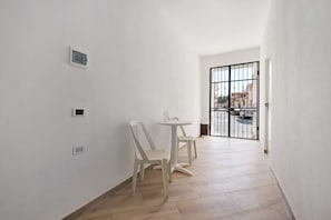 Un pasillo estrecho con paredes blancas, una pequeña mesa con sillas y una reja que da al exterior.