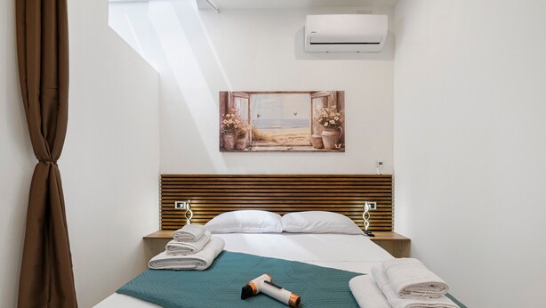 A hotel room with a bed, a teal blanket, a rolled towel, a portable heater, and a painting of a beach scene.