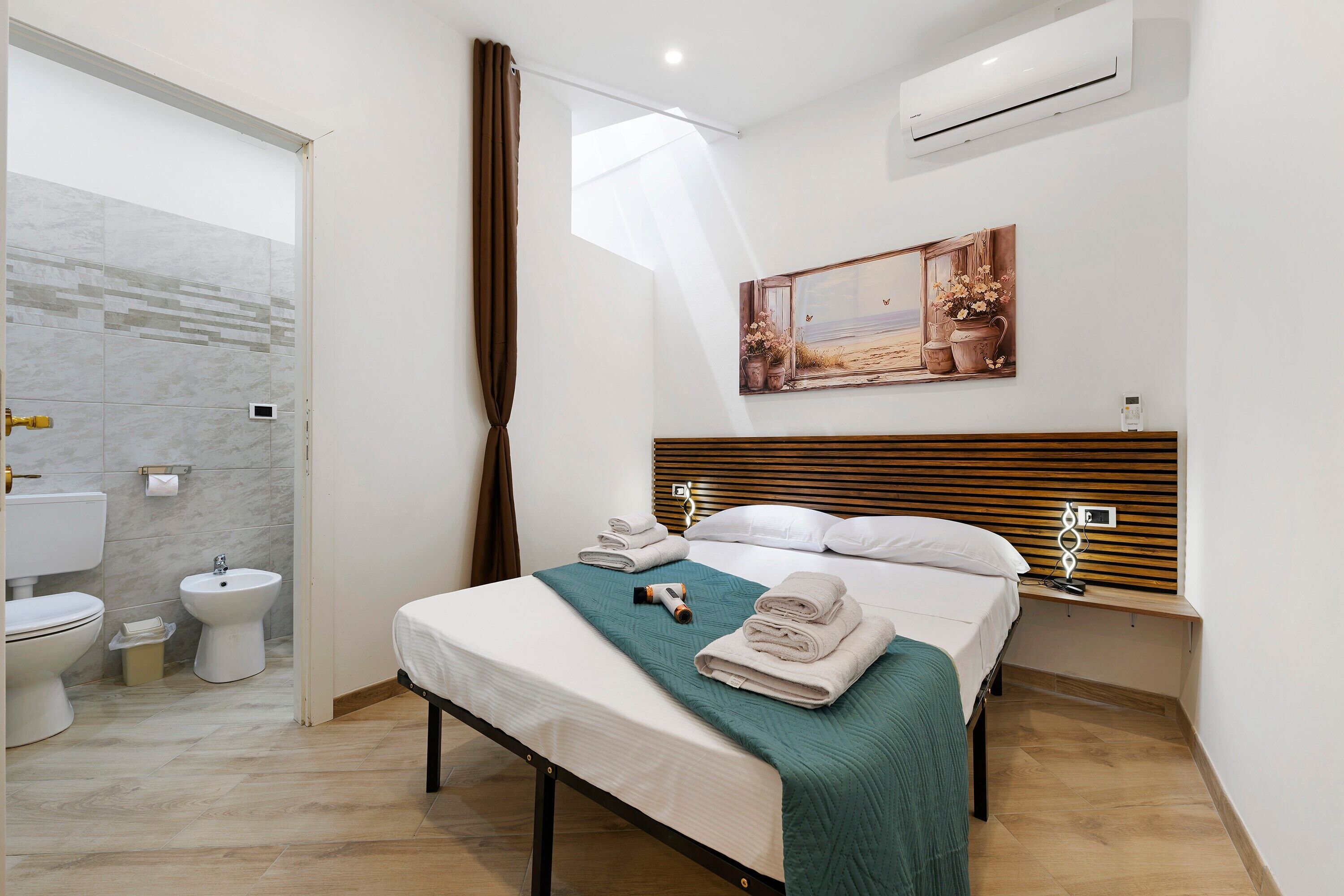 A hotel room with a bed, bedside table, wall-mounted air conditioner, and a painting.