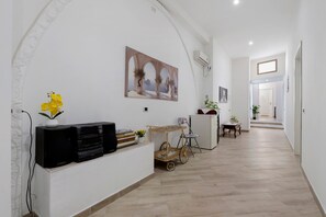 Un pasillo con pared blanca, un cuadro de una escena costera, una mesita con una planta y un mueble para televisor con un altavoz.