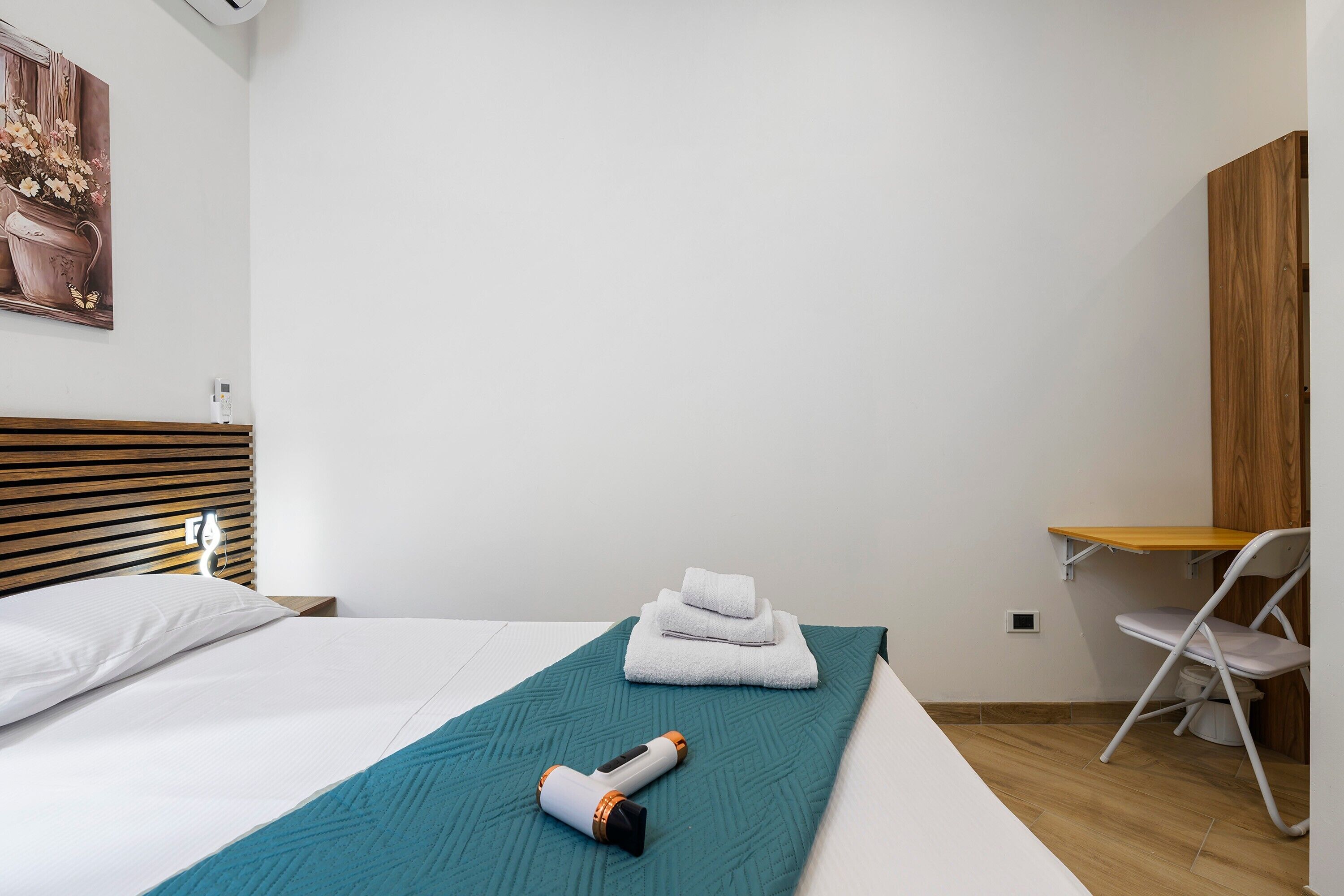 A hotel room with a bed, a teal blanket, folded towels, a hairdryer, a wooden headboard, a white chair, and a wooden desk.