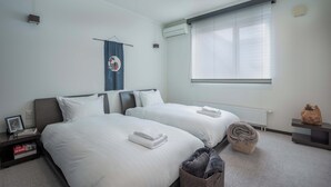 A hotel room with two beds, a radiator, a window with blinds, a wall-mounted air conditioner, and a small table with a decorative item.