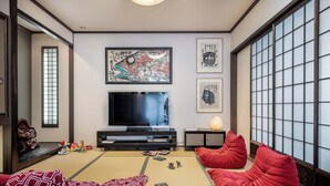 A traditional Japanese-style living room with tatami flooring, a flat-screen TV, red bean bag chairs, and artwork on the walls.