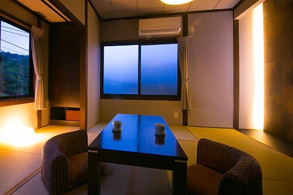 A traditional Japanese room with tatami flooring, a low table, cushions, and a window with a view.