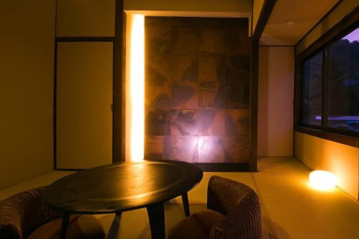 A room with tatami flooring, a low table, cushioned seating, a stone wall, and a view through a window.