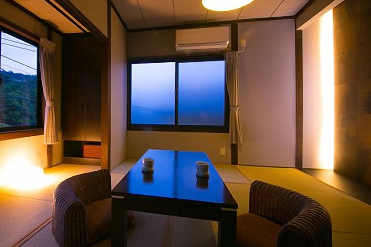 A traditional Japanese room with tatami flooring, a low table, cushions, and a window with a view.