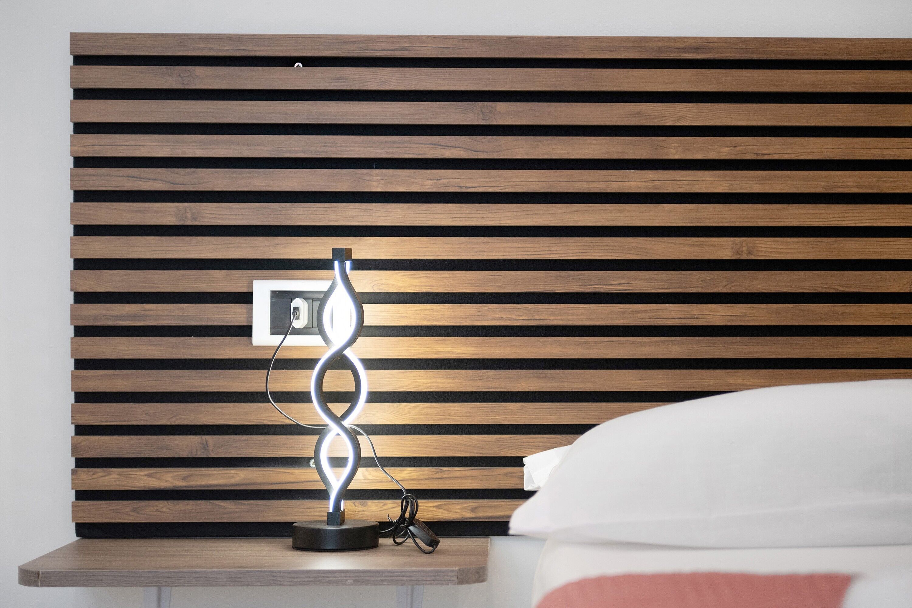 Ein Bett mit Holz-Kopfschild, ein Nachttisch mit einer modernen Lampe und eine Steckdose.