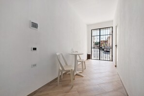 Un couloir étroit aux murs blancs, une petite table avec des chaises et un portail grillagé donnant sur l’extérieur.
