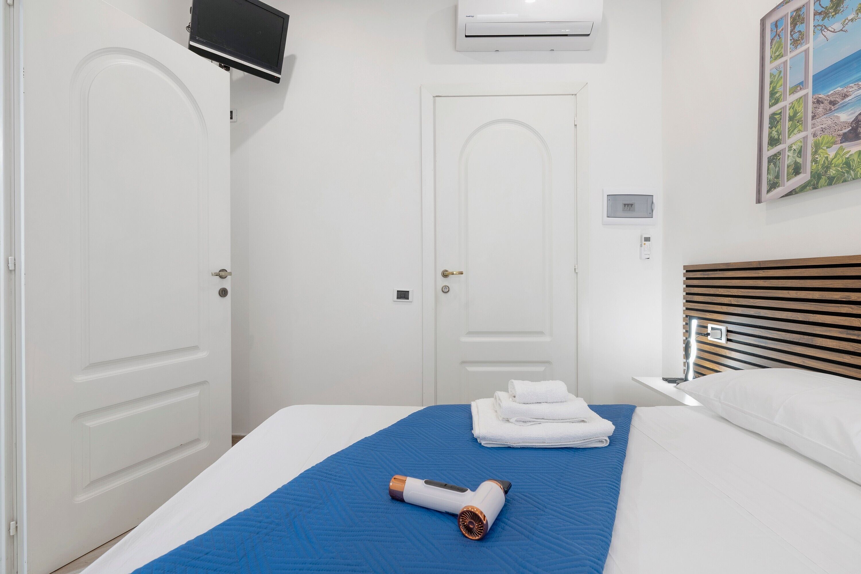 A hotel room with a bed, a blue bedspread, a rolled-up towel, a hairdryer, and a painting of a beach scene.
