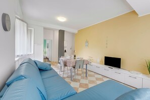 Ein Wohnzimmer mit einer Couch, einem Esstisch mit Stühlen, einem Flachbildfernseher und einer gelben Wand.