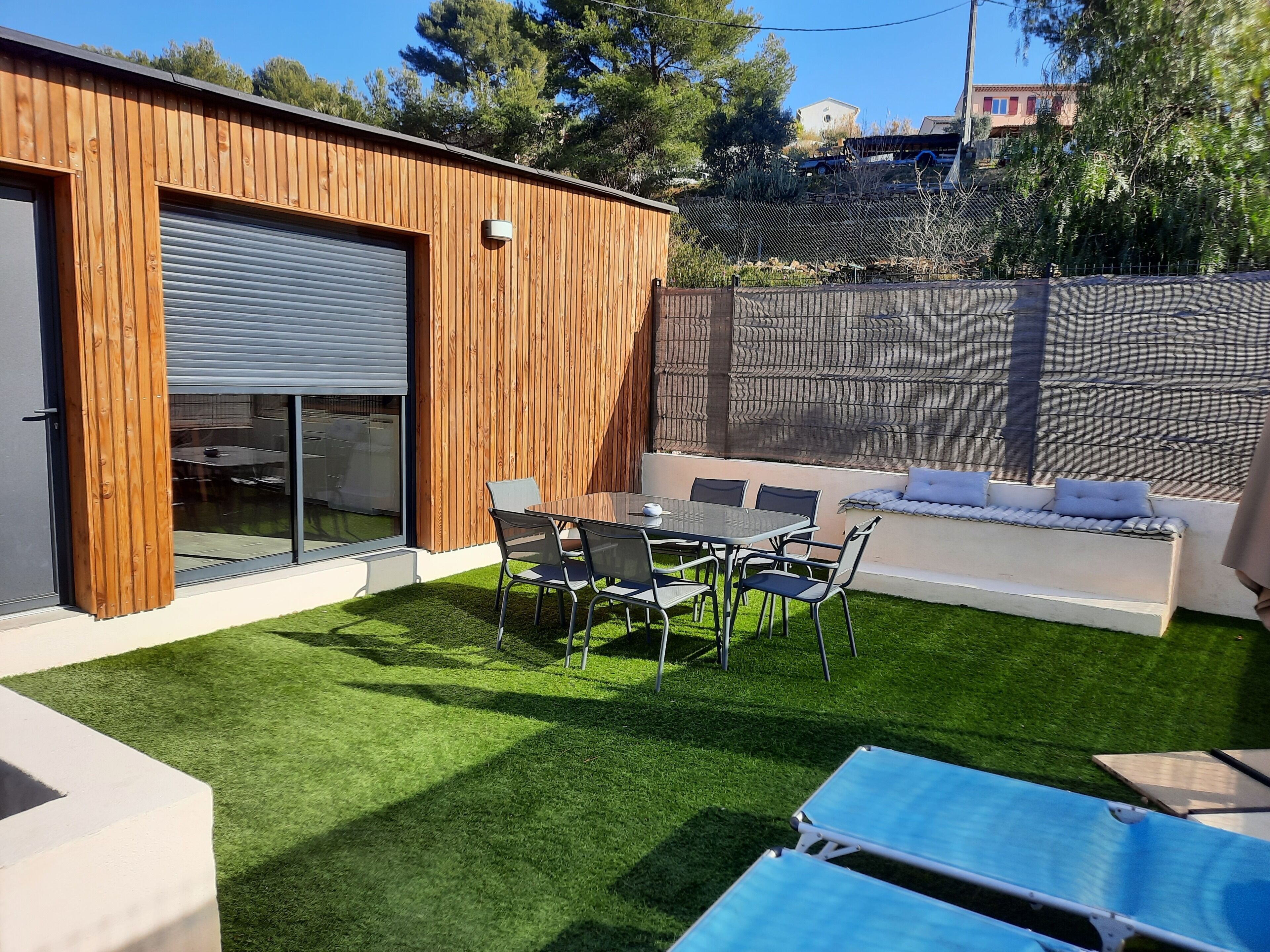 Un patio ext érieur moderne avec un mur en bois, une porte coulissante en verre, une table à manger avec des chaises et un coin salon avec des coussins.