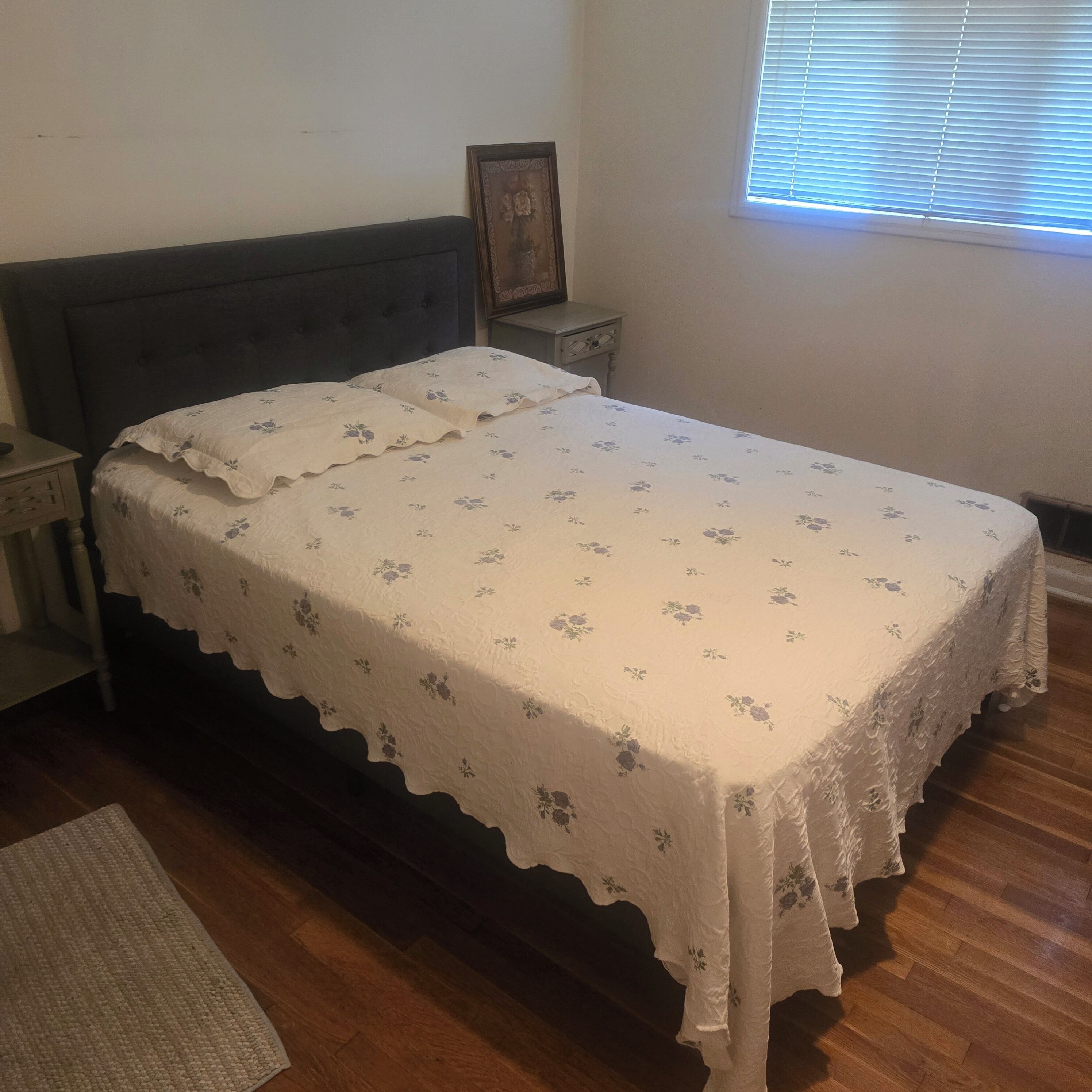 Ein Bett mit floraler Tagesdecke, ein Nachttisch mit Lampe und ein Fenster mit Jalousien.