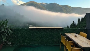 Un patio con mesa y sillas, un seto verde y vistas a montañas parcialmente cubiertas por la niebla.