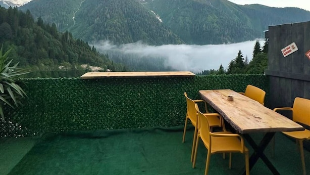 Una terraza con una mesa de madera y sillas amarillas, con vistas a un valle con montañas y un lago.