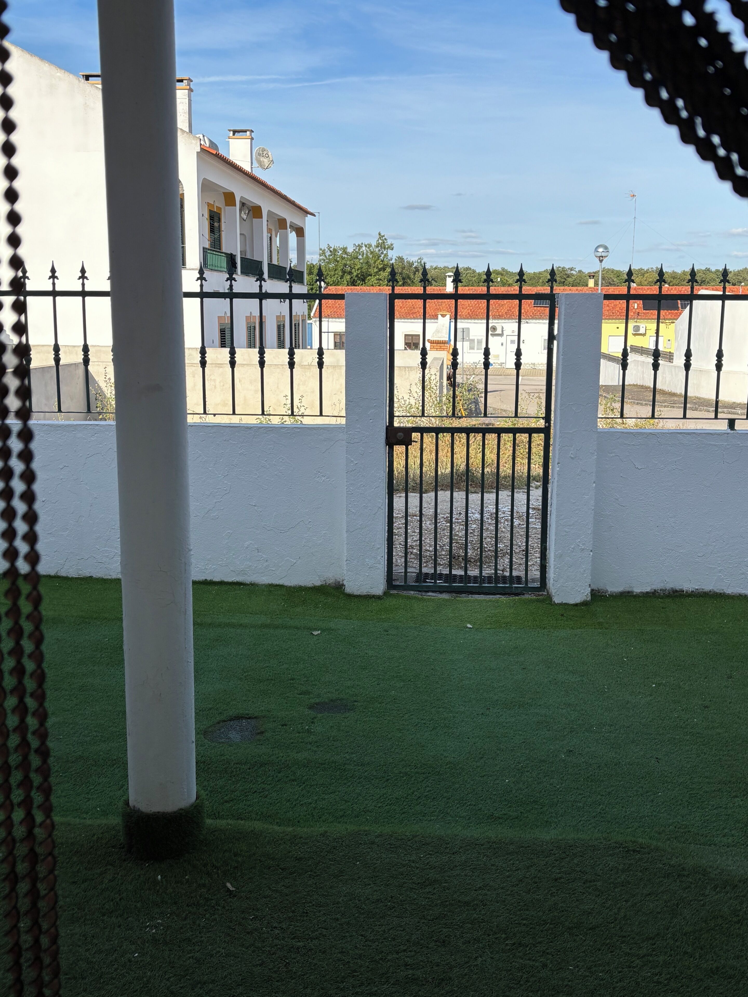 A gated area with a white wall, a metal gate, and a building with a red roof in the background.