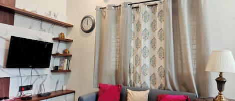 Una sala de estar con televisor de pantalla plana, un reloj, un sofá con cojines estampados, una mesita de centro y una lámpara.