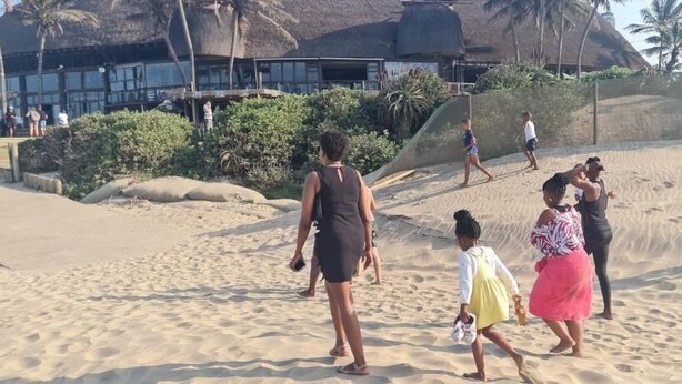 A sandy beach with people walking, palm trees, and a building with a thatched roof in the background. - Yoxall Overnight stays (Durban)
