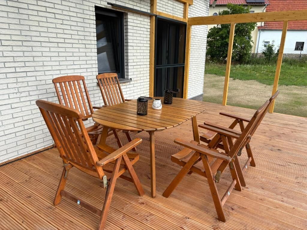 Een houten terras buiten, met een tafel, stoelen, een bakstenen muur en een raam.
