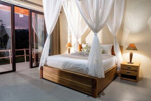 A bedroom with a wooden bed, white bedding, a canopy, two bedside tables with lamps, and a sliding glass door leading to an outdoor area. - Cassowary Villas Sidemen by AGATA (Karangasem)