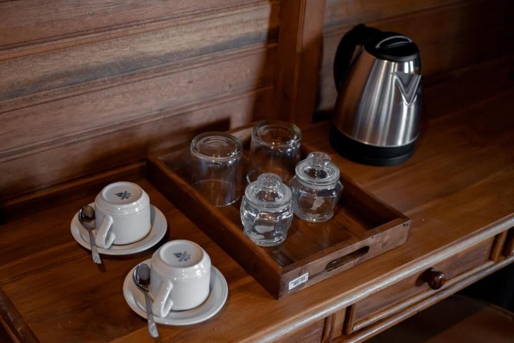 Una bandeja de madera con dos tazas y platillos blancos, una cuchara y tres recipientes de vidrio sobre una superficie de madera.