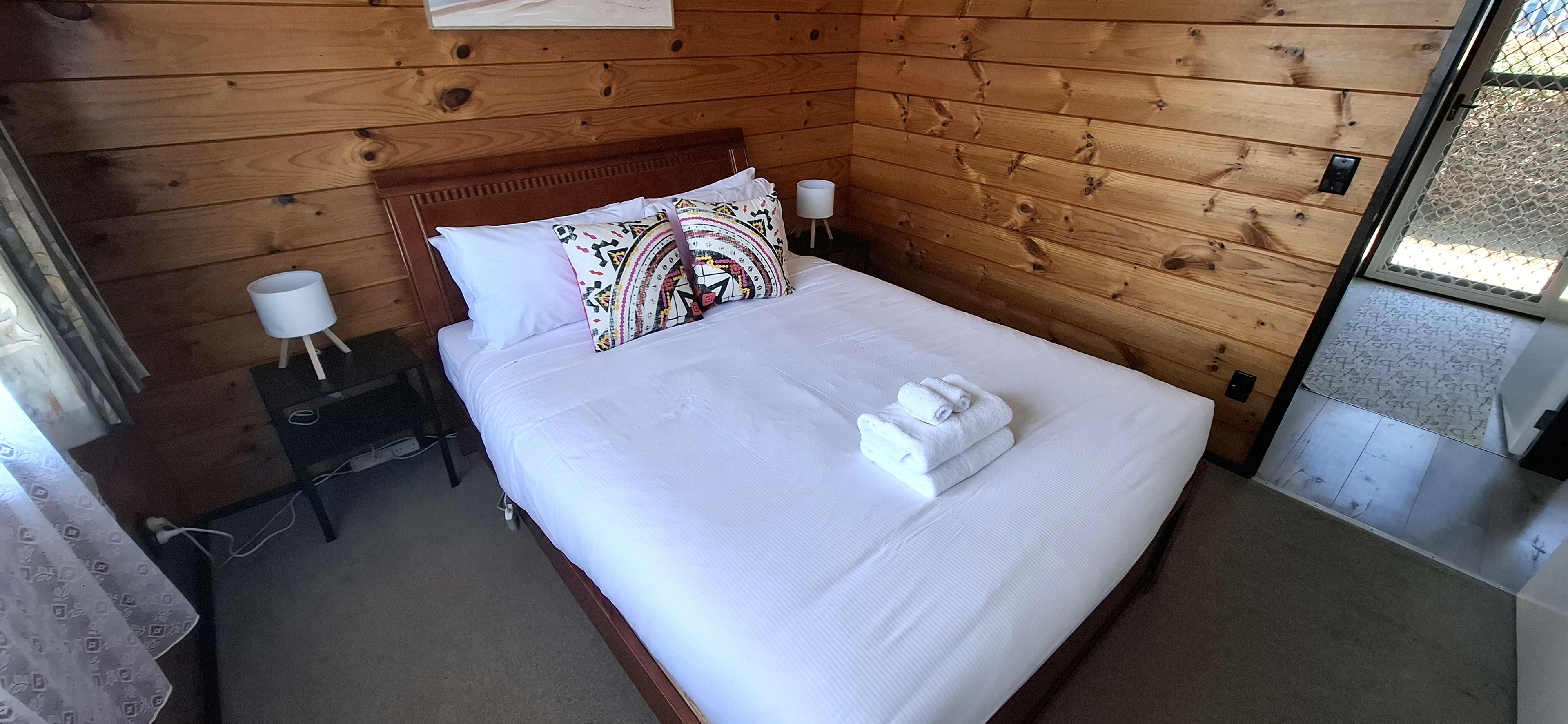 Une pièce aux murs lambrissés, avec un lit, du linge de lit blanc, un coussin à motifs, une petite table de chevet avec une lampe et une prise électrique.