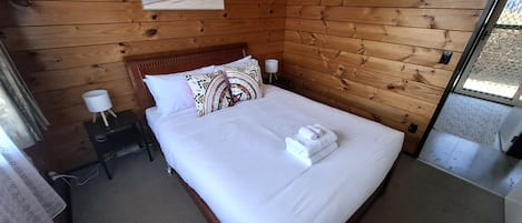 Habitación con paneles de madera, una cama, ropa de cama blanca, un cojín estampado, una mesita de noche pequeña con una lámpara y un enchufe.