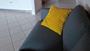 Un salon avec un canapé, un coussin jaune, un coin cuisine avec des placards et un escalier en bois.