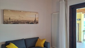 Une pièce avec un canapé bleu, un tableau représentant un paysage urbain, un radiateur et une vue par la fenêtre.