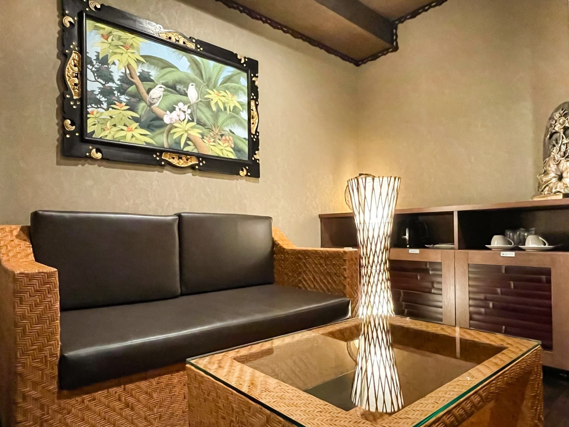 A room with a black leather sofa, a glass coffee table, a decorative vase, a wall-mounted painting, and a wooden cabinet with dishes.