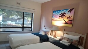 A hotel room with two beds, a nightstand with a lamp, a painting of a sunset, and a window with a view of trees.