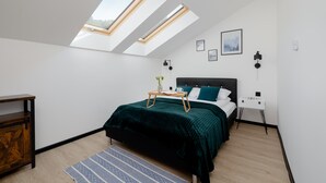 Une chambre avec une lucarne, un lit recouvert d’une couvre-lit vert foncé, une table de chevet avec une lampe et une petite table de nuit blanche.