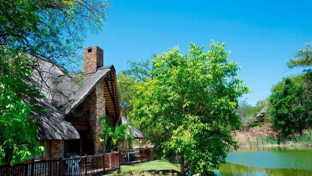 A rustic lodge with a stone chimney, a fenced deck, and a pond.