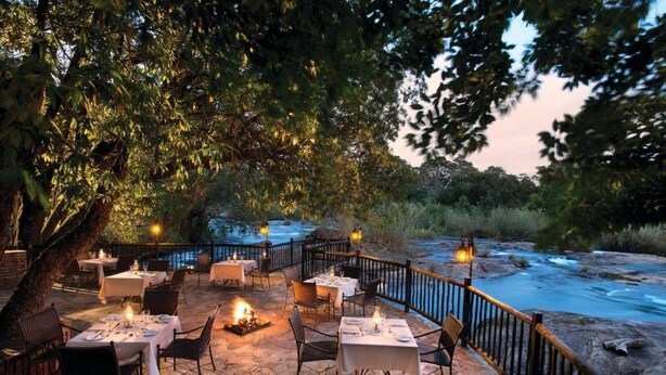 Un espacio de comedor al aire libre con mesas y sillas, un hogar a leña y vistas de un río.