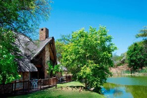 A rustic lodge with a stone chimney, a fenced deck, and a pond. - Kruger Park Lodge 2-bedroom WBT Rentals (Ehlanzeni District Municipality)