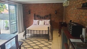 Une chambre avec un mur en briques, un lit avec une literie à motifs floraux, une fenêtre donnant sur un paysage agréable, une petite table et un coin cuisine.
