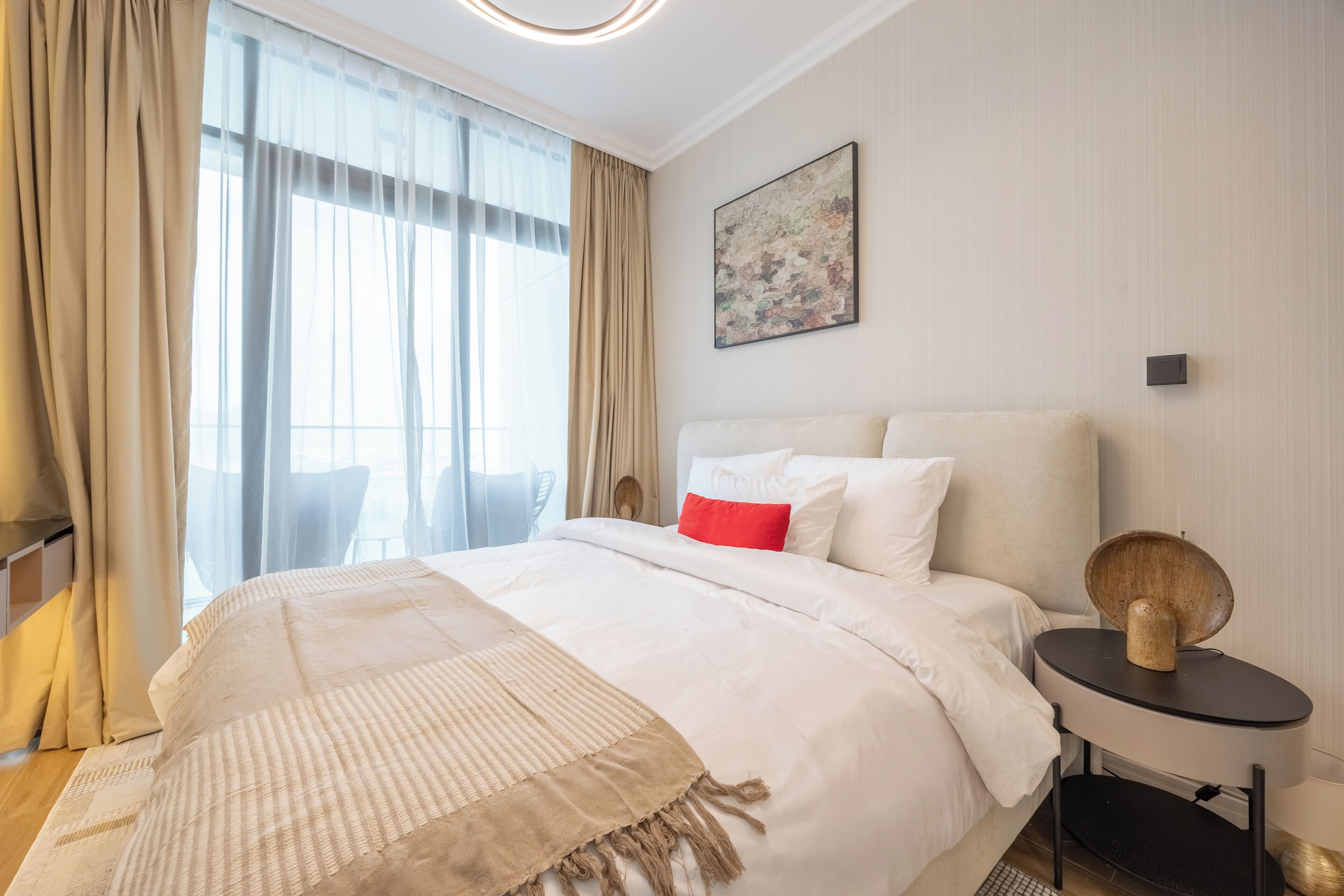 Ein Hotelzimmer mit einem großen Bett, einem Nachttisch mit Lampe, einem Fenster mit Vorhängen und einem Wandbild.