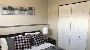 Un lit bien fait, recouvert d’une couverture grise, avec deux coussins à carreaux noirs et blancs, une lampe de chevet et une étagère à plantes fixée au mur.