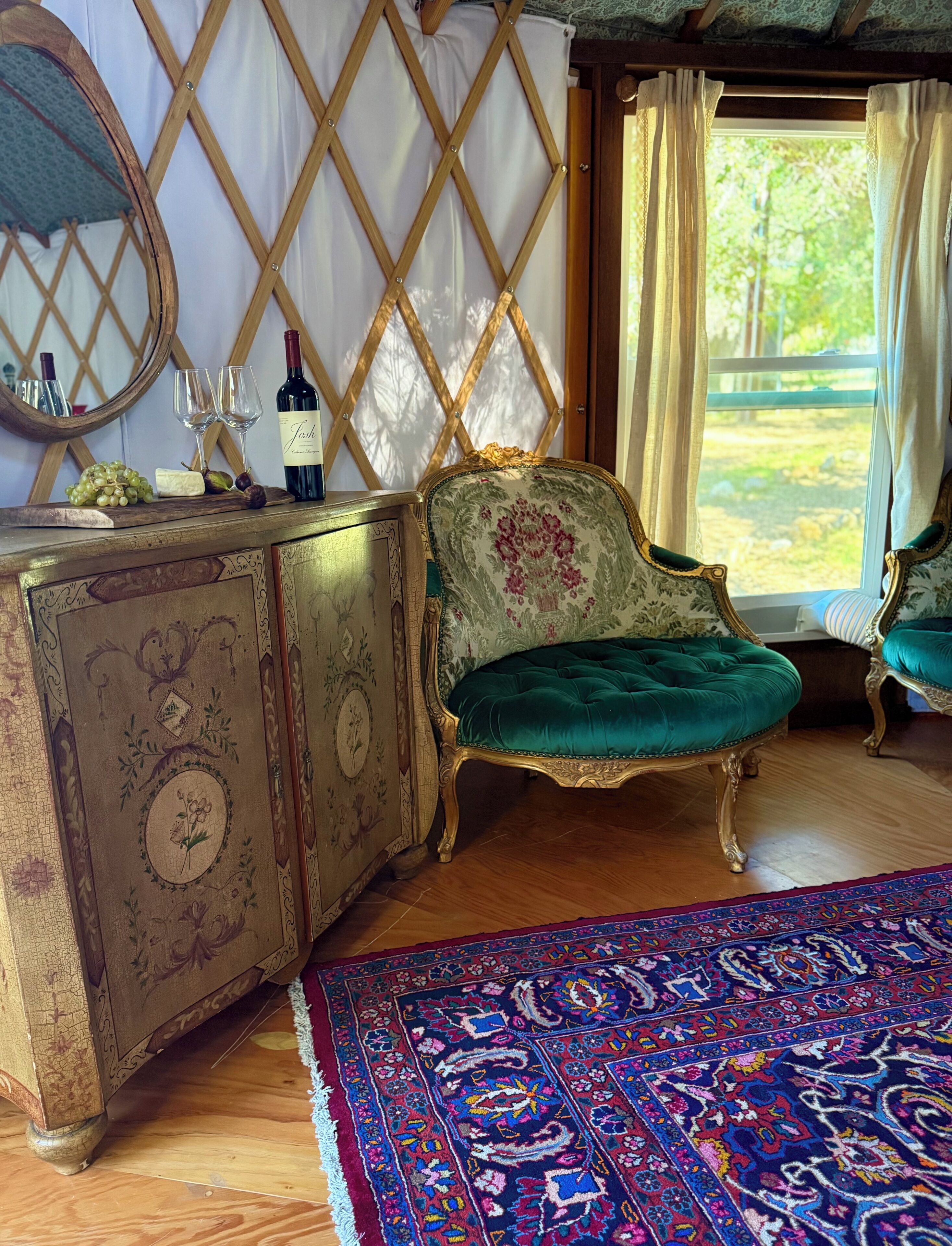 A room with a vintage-style wooden cabinet, a green upholstered chair, a patterned rug, and a window with curtains.