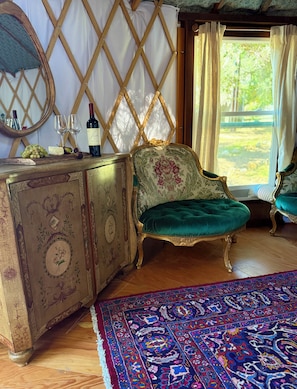 A room with a vintage-style wooden cabinet, a green upholstered chair, a patterned rug, and a window with curtains.