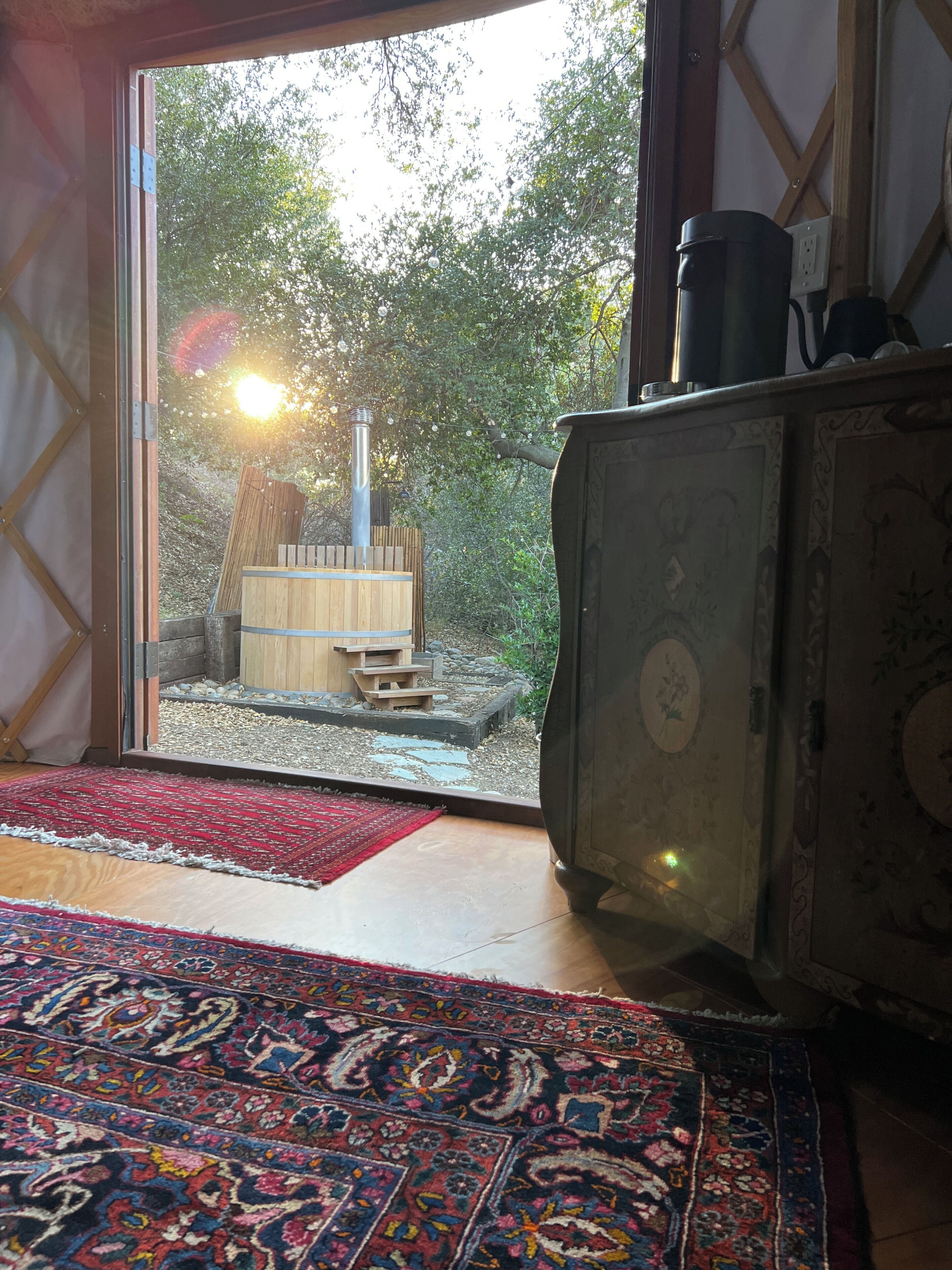 Une pièce dotée d’une grande porte-fenêtre coulissante qui donne sur un espace extérieur agrémenté d’une structure en bois et d’une cheminée. Le sol est recouvert d’un tapis à motifs et la pièce contient un meuble au design décoratif.
