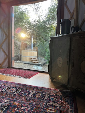 Una habitación con un amplio ventanal corredero que da a un espacio exterior con una estructura de madera y una chimenea. Hay una alfombra estampada en el suelo y un mueble con detalles decorativos.